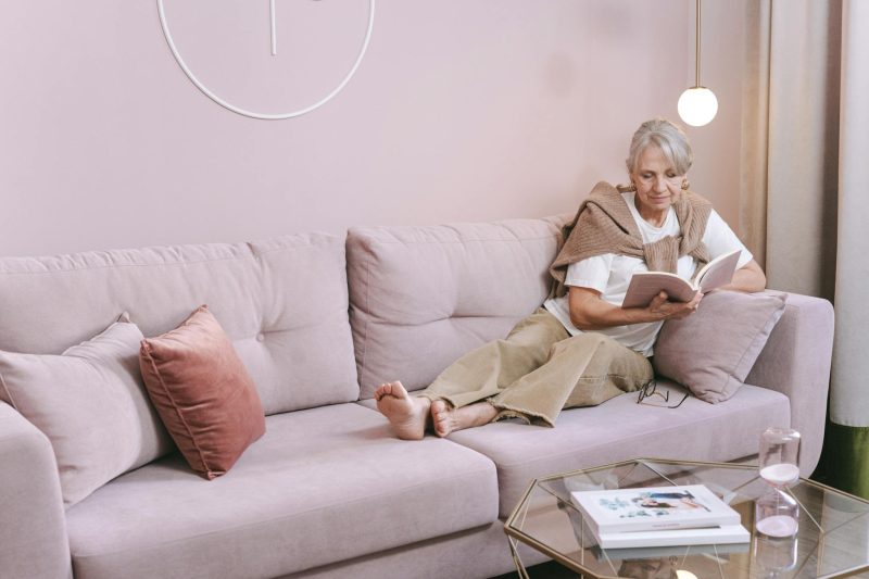 Photo of an Elderly Woman Reading a Book while Sitting on a Sofa