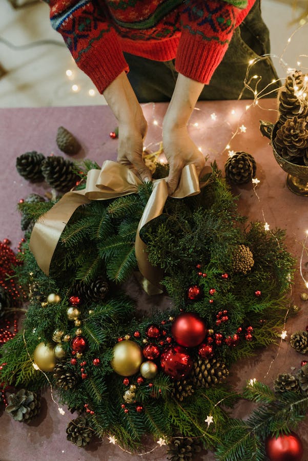 A close-up of hands arranging a Christmas wreath with ornaments and ribbon on a decorated table.