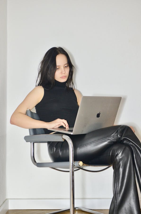 Young woman sitting in an armchair, using laptop, focused and thoughtful in a modern indoor setting.