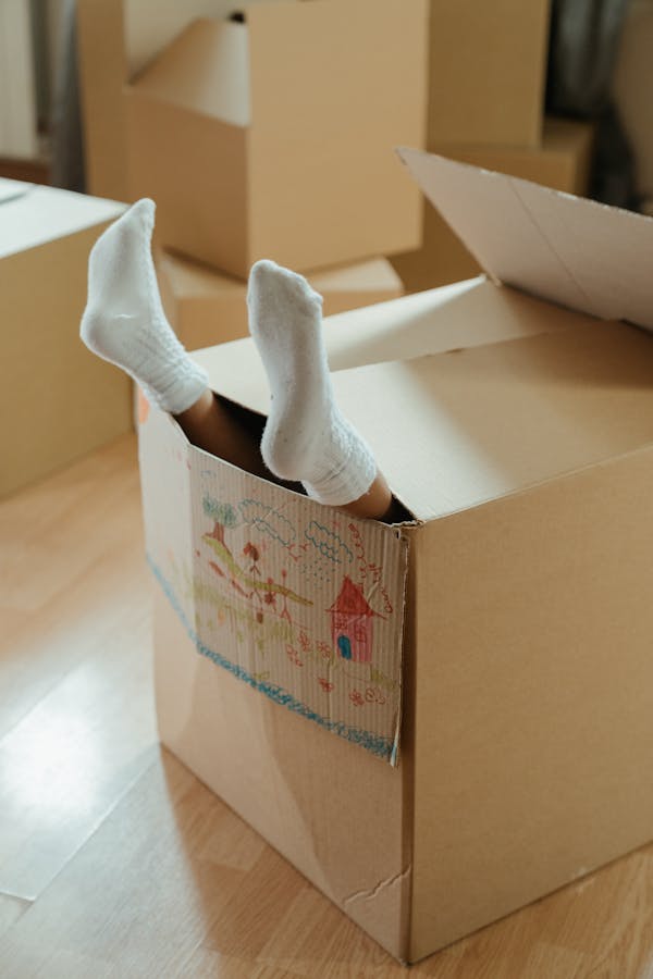 A child's feet in summer socks sticking out of a cardboard box inside a home, symbolizing fun and imagination during moving.