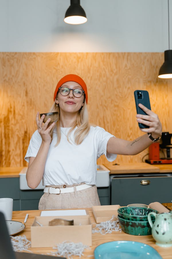A woman in a beanie and glasses smiles while taking a selfie holding a bowl in a modern kitchen.