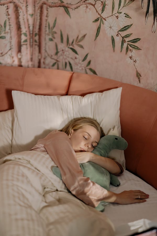 A woman in pajamas peacefully sleeping in bed, cuddling a plush toy, with a floral-themed wall behind.