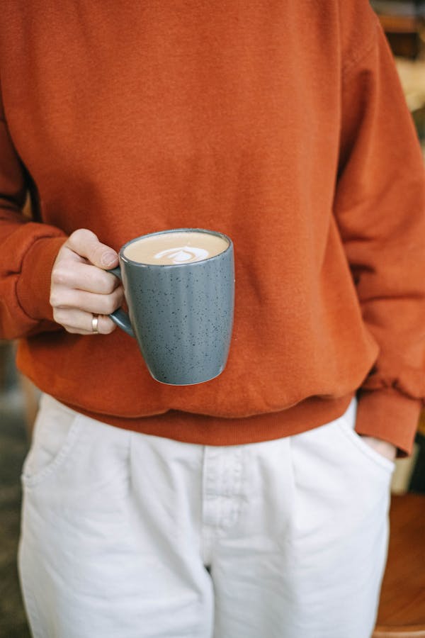 Person in orange sweater enjoys a cozy latte from a ceramic mug, perfect for chilly days.