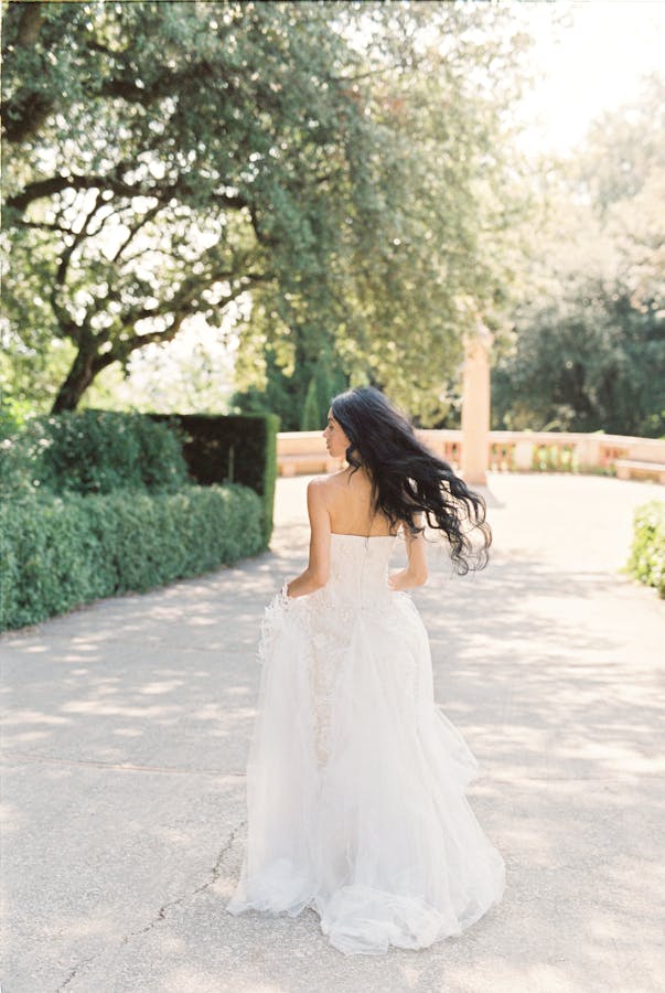A bride with flowing dark hair walks outdoors in a beautiful white gown, surrounded by lush greenery.