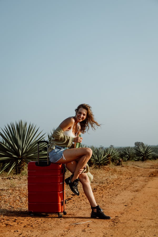 Stylish woman seated on red suitcase outdoors, embracing travel vibes.