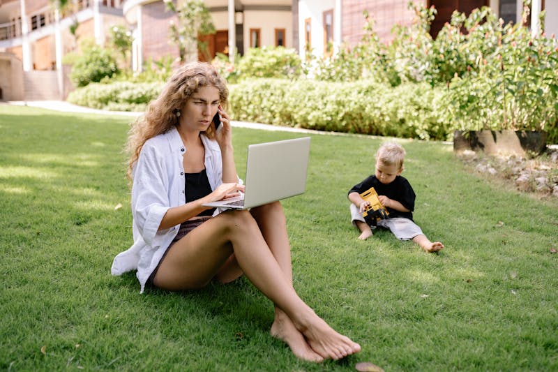 Woman working remotely on a laptop outdoors while her child plays nearby in the grass. smart tools for moms