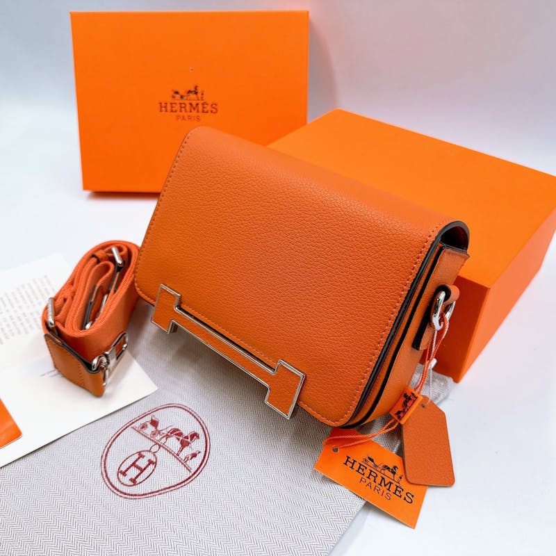 Close-up of an orange Hermes leather handbag with box and tag, showcasing luxury fashion.
