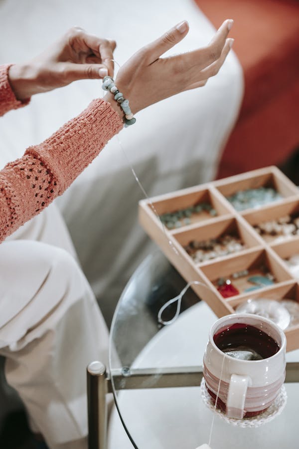 A person arranges beads for a handmade bracelet with a cup of tea nearby.