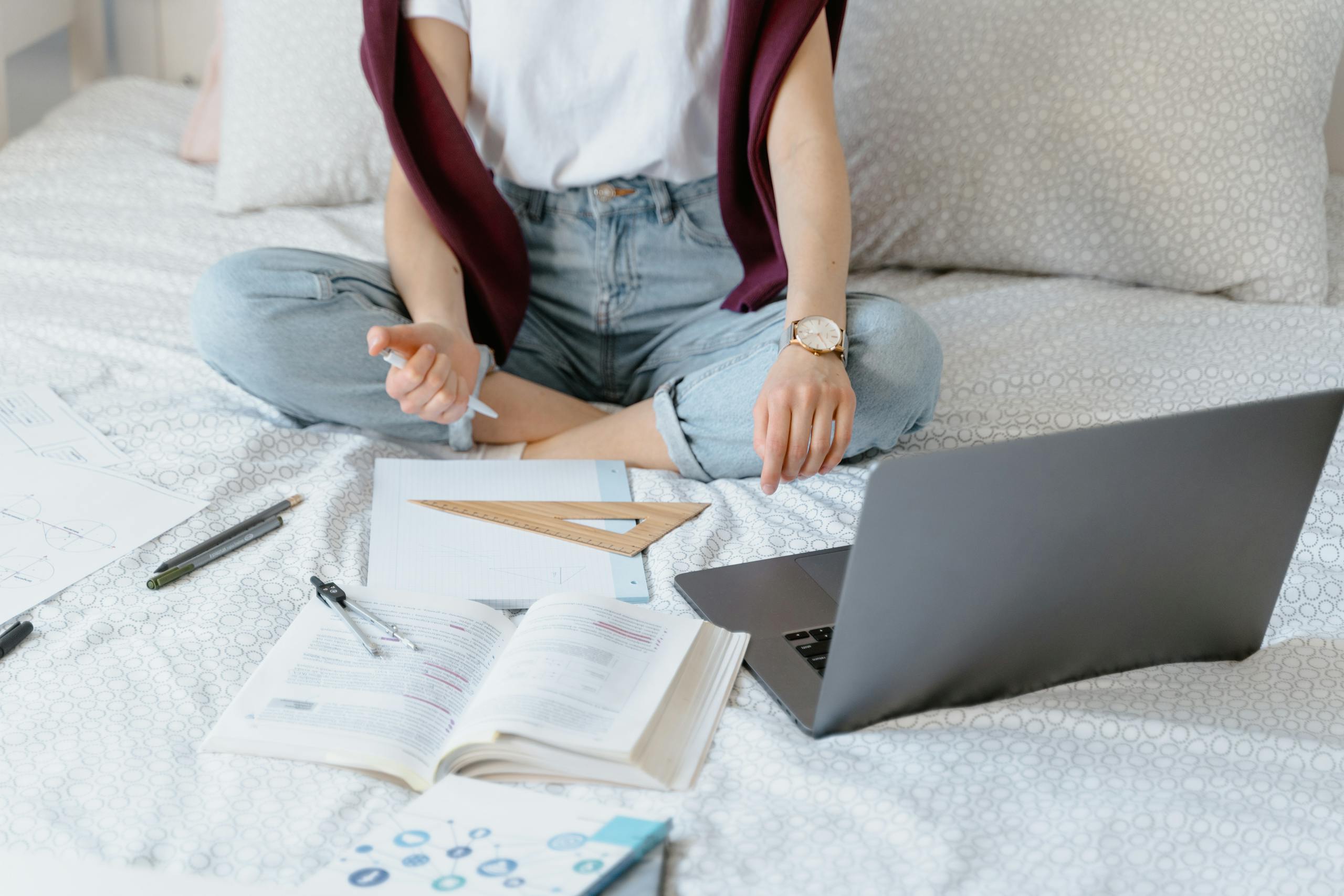 A young woman sitting on a bed using a laptop for remote learning, books and papers scattered around.