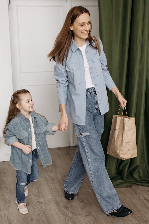 A happy mom and daughter holding hands, wearing denim, and smiling while walking indoors.